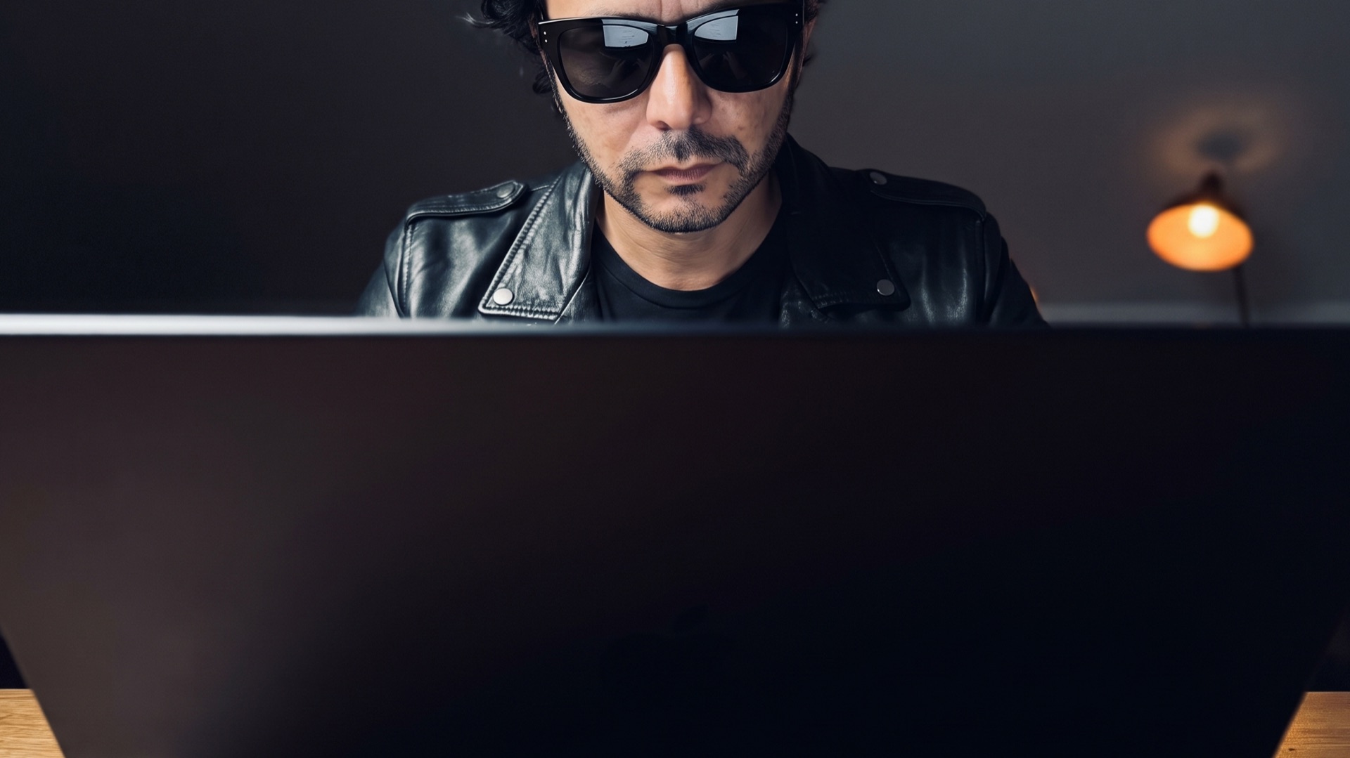 Javier Amaya at work behind a black MacBook Pro, seen from the back of the laptop, in sunglasses and a black leather jacket.